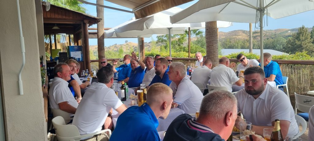 Group of golfers sitting outside under parasols enjoying some beverages.