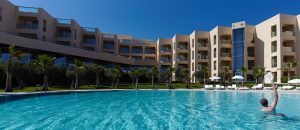 The swimming pool at the Sao Rafael Suites with the hotel in the background and a swimmer playing padel in the pool.