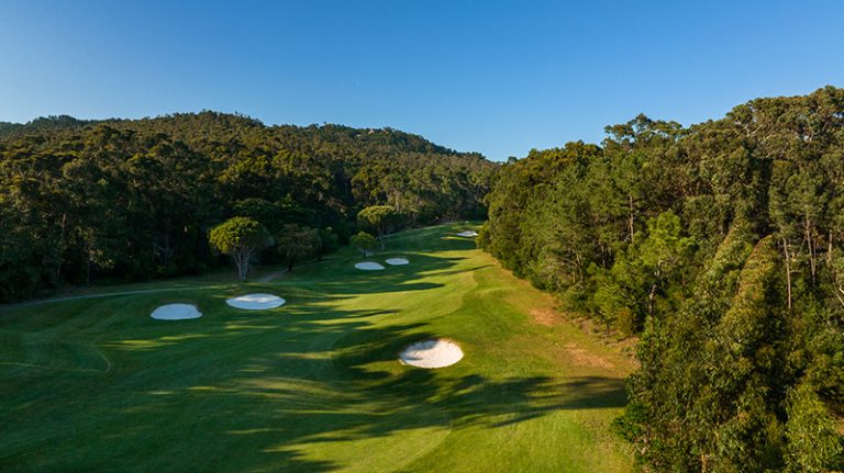 A panoramic view of the Penha Longa Atlantico golf course, featuring a green fairway sloping towards the ocean, surrounded by trees and clear skies.