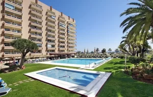 Hotel PYR in Fuengirola, featuring a spacious outdoor pool and a smaller children's pool, surrounded by sun loungers and well-maintained green lawns, with palm trees and a multi-story building in the background.