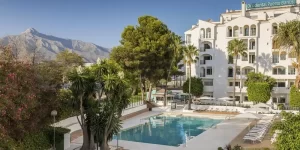 Elegant exterior of Occidental Puerto Banus hotel, featuring a spacious outdoor pool area surrounded by palm trees, with a backdrop of mountains under a clear blue sky.