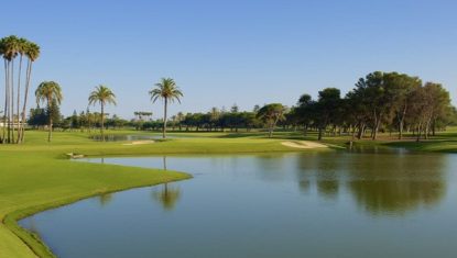 Scenic view of the Real Club de Golf Sotogrande featuring lush greenery, palm trees, and a calm water hazard under a clear blue sky.